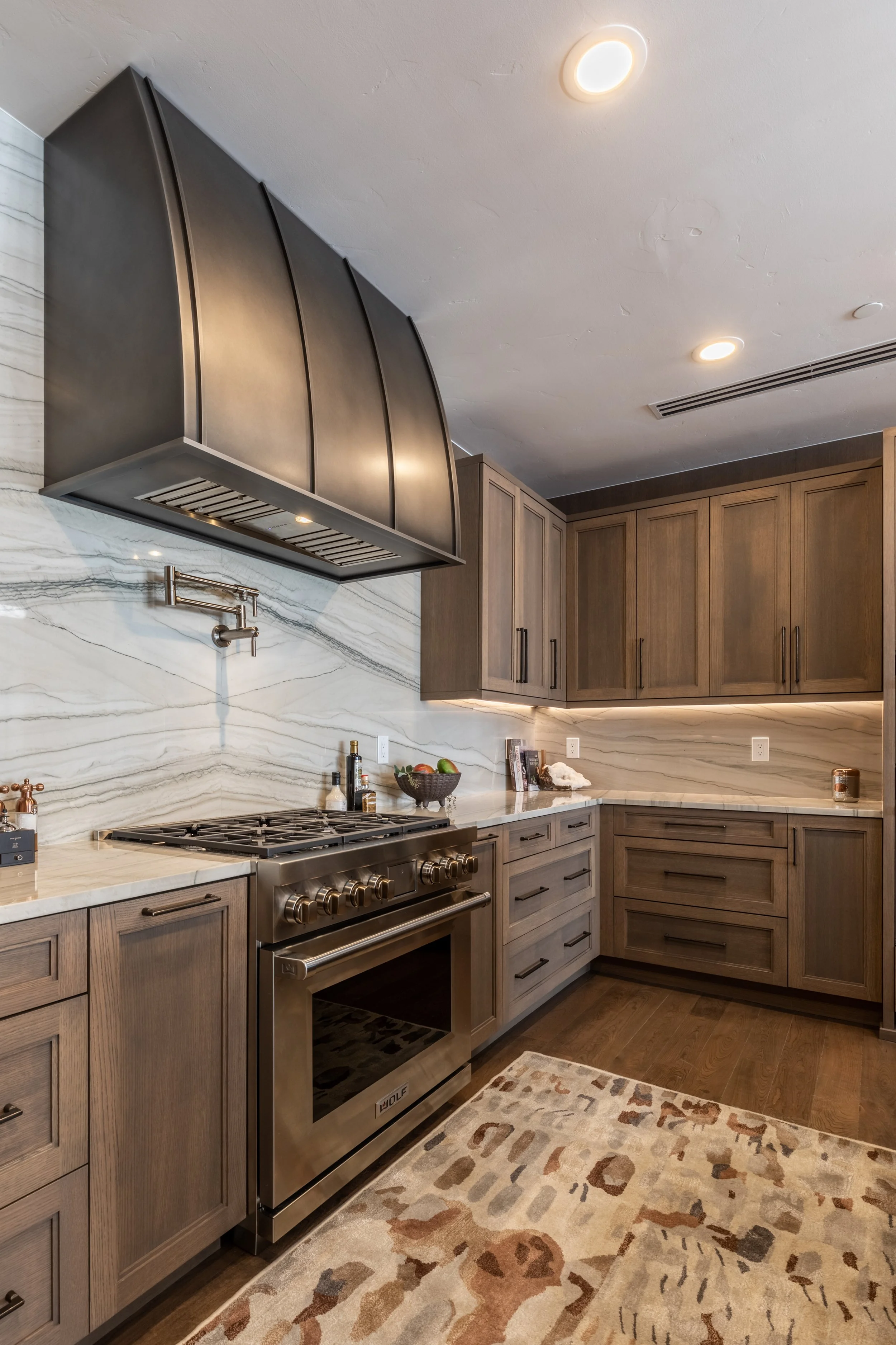 A modern kitchen with wooden cabinets, a marble countertop, stainless steel appliances, a stove, a grey range hood, a colorful area rug, and decorative items on the counter.