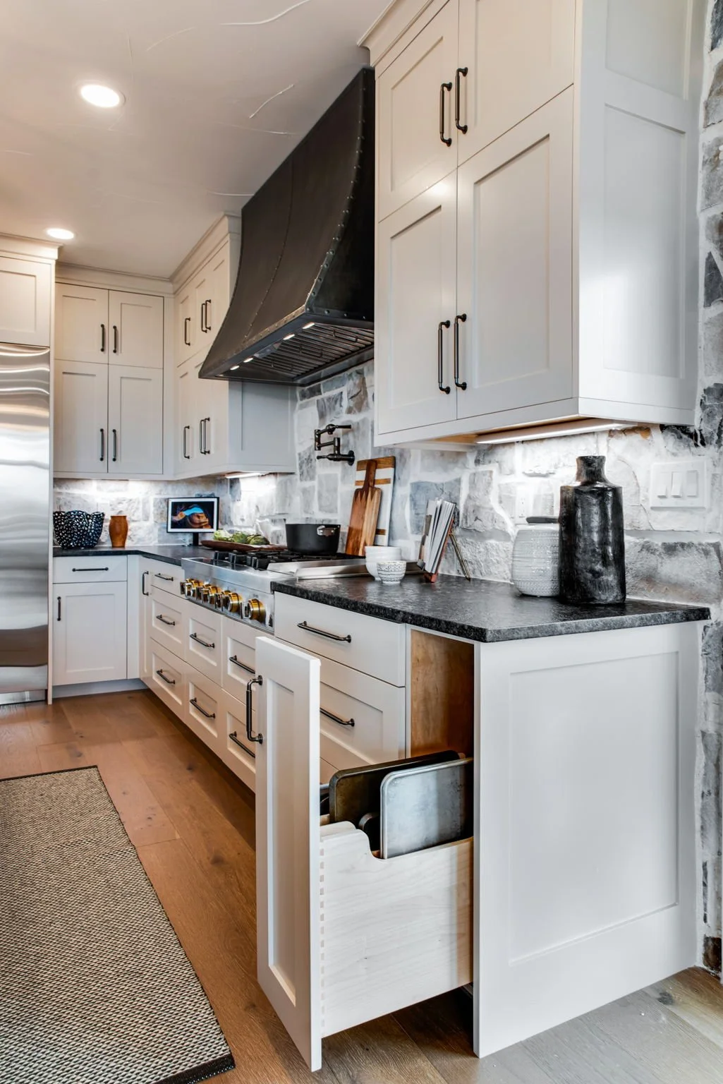 Modern kitchen with white cabinets, black handles, a black range hood, stone backsplash, and wooden flooring. Open drawer shows kitchen utensils inside.
