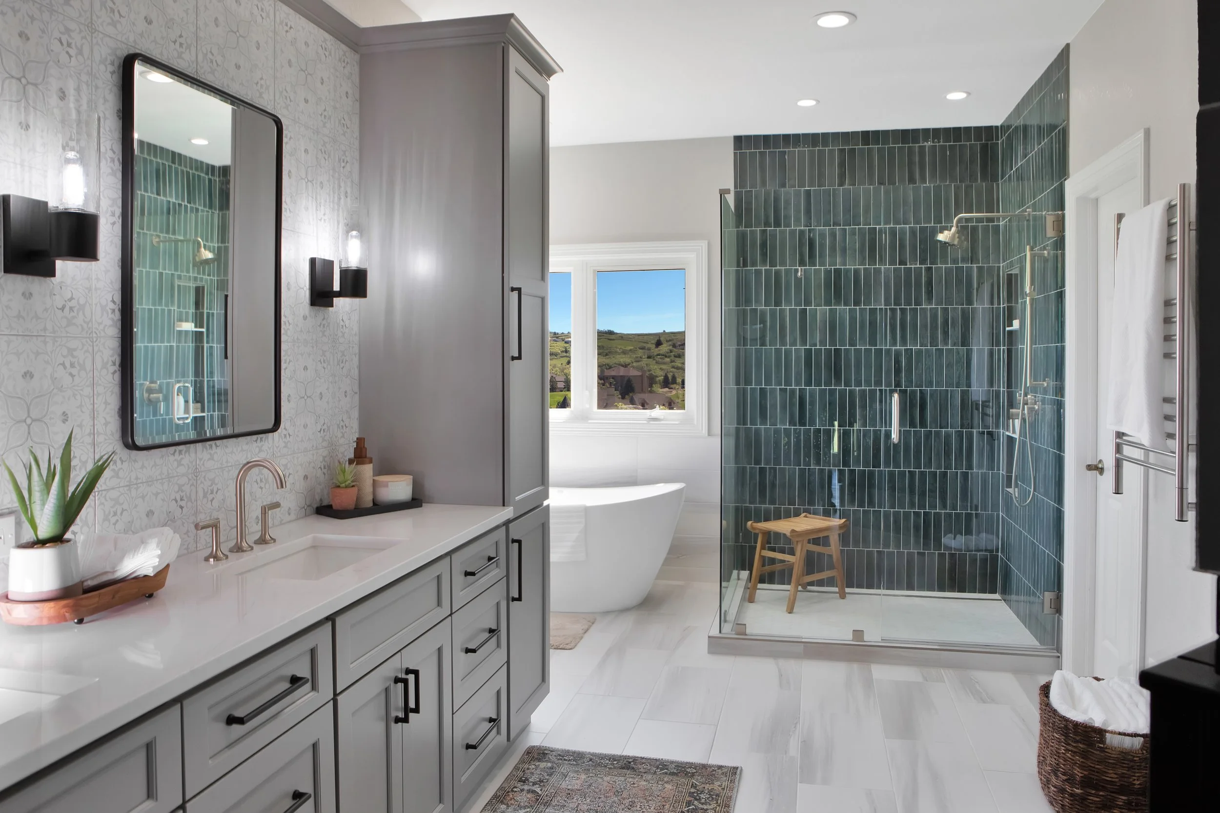 Modern bathroom with a light gray vanity, black hardware, a large frameless mirror, two wall sconces, a white bathtub near a window, and a walk-in shower with dark green tiles and a wooden bench.