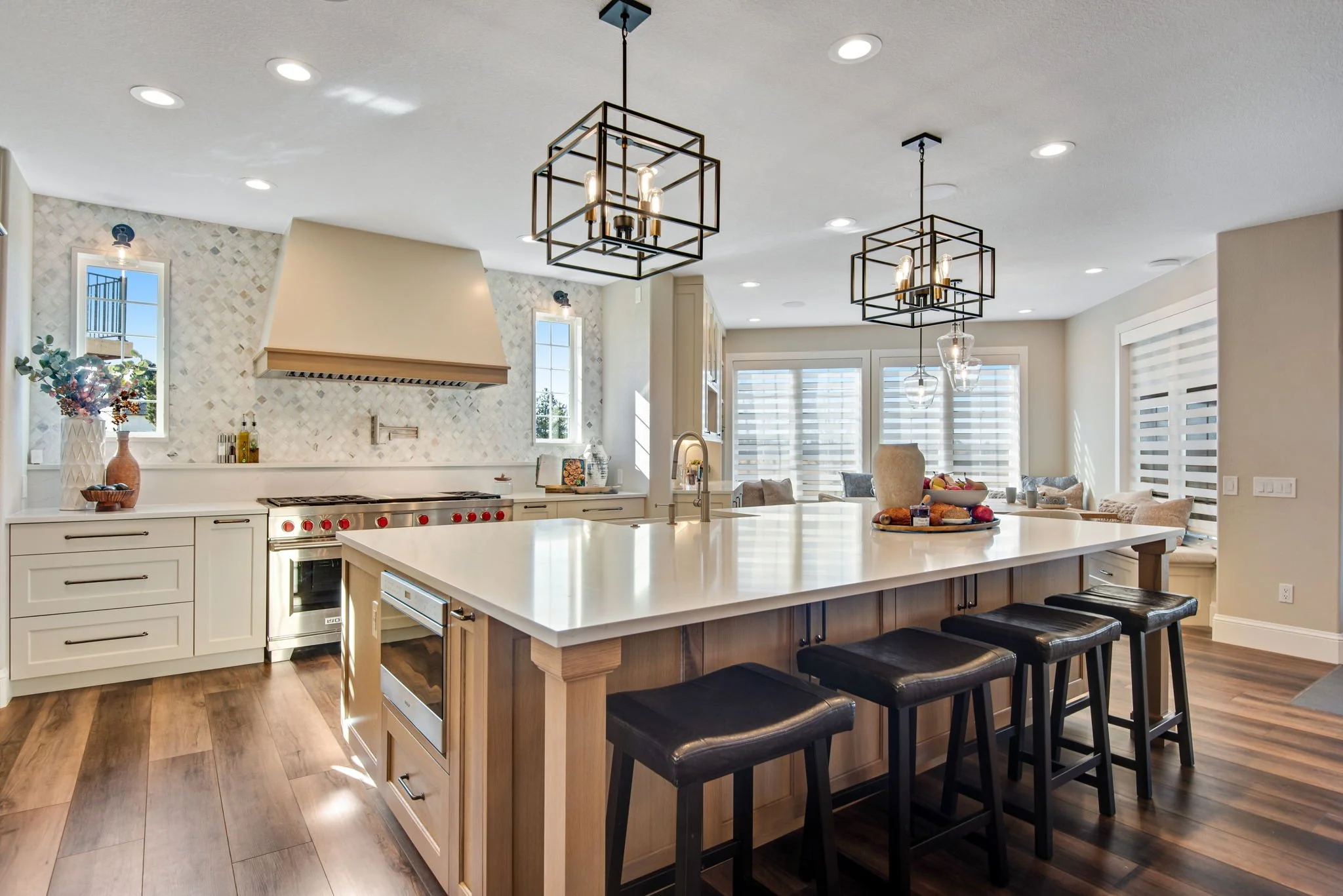 Modern kitchen with white cabinetry, a large island with a white countertop and black bar stools, a stainless steel stove, decorative pendant lights, and a sitting area with patio doors and large windows.
