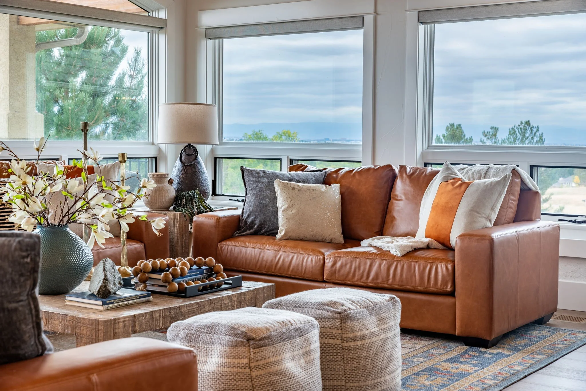 Living room with large windows, brown leather sofa with cushions, wooden coffee table with decorative items, cream poufs, and a patterned rug.